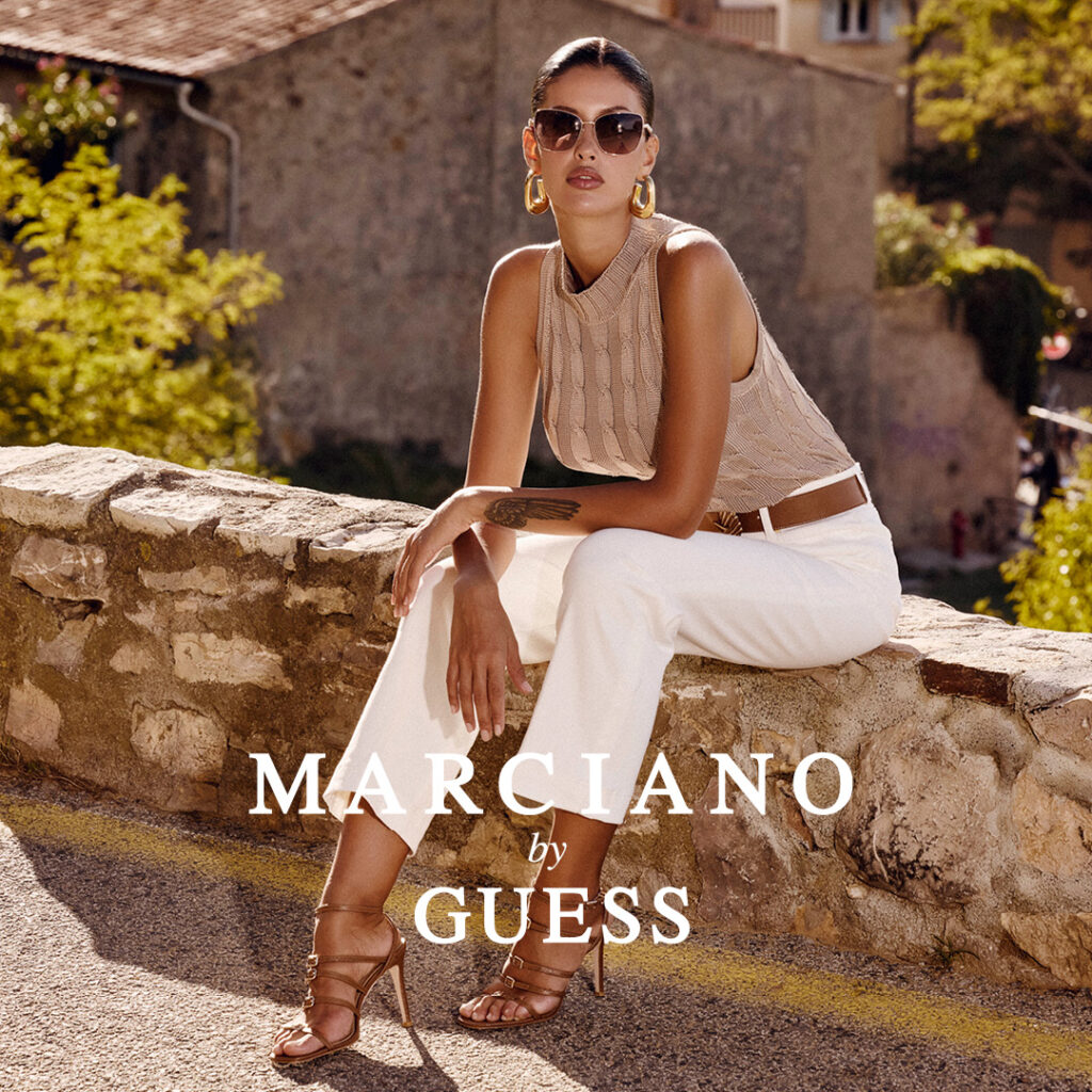 Kobieta w letniej stylizacji siedząca na murku, w tle kamienne budynki.